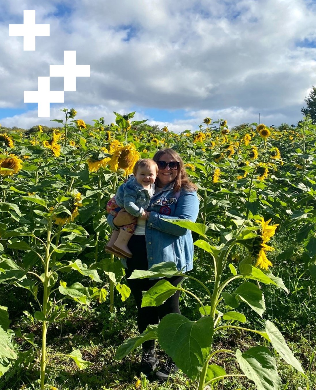 A photograph of Leigh Miele, Training Co-ordinator in Employability at UHI Moray. She is standing in a field of sunflowers holding her young daughter. She has long dark brown hair, is wearing a light blue denim shirt, black trousers and sunglasses, smiling at the camera.