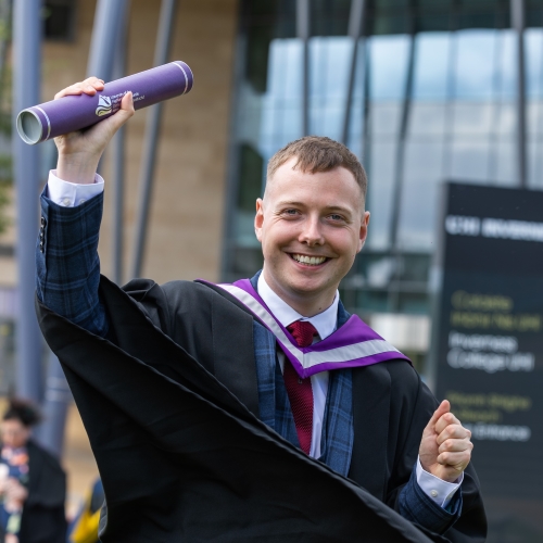 George Gunn in his graduation gown, holding up his scroll and smiling