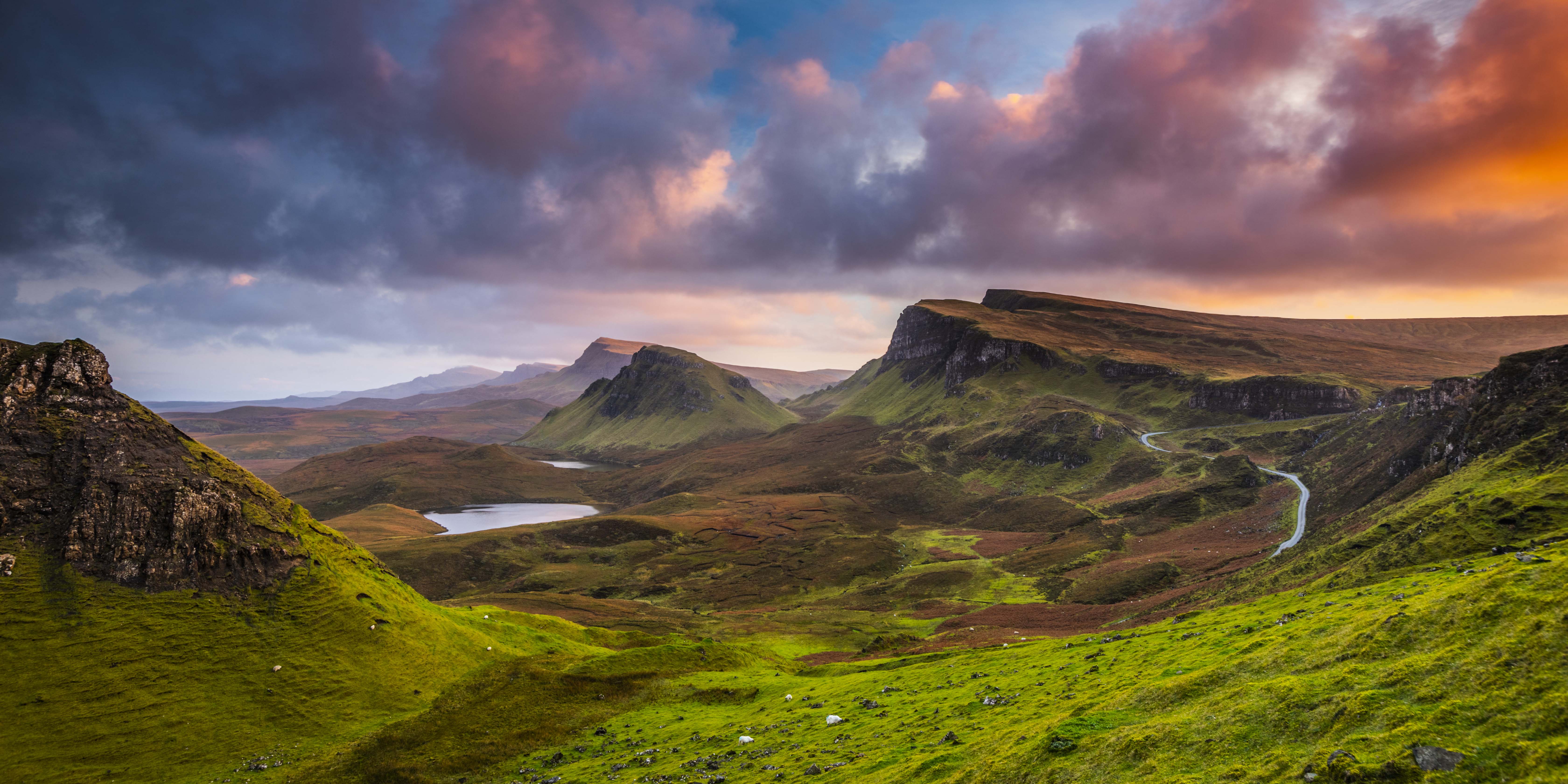 This image depicts a stunning natural landscape featuring rolling green hills, dramatic cliffs, and a winding road leading into the distance. The scene is bathed in vibrant colors, with a dramatic sky above transitioning from deep blues to fiery oranges and reds, indicating either sunrise or sunset.