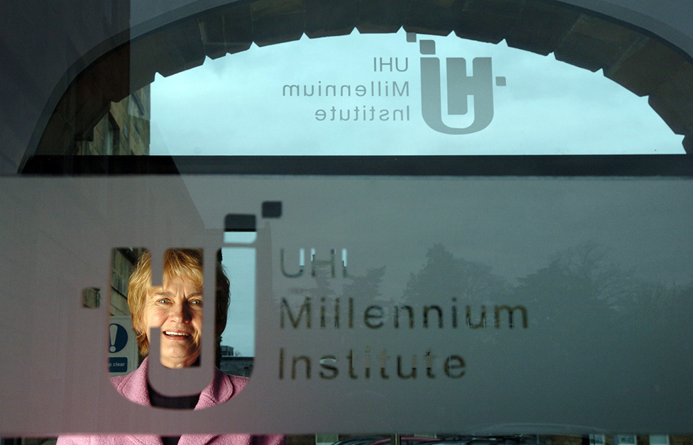 Dr Val MacIver OBE standing with the UHI Millenium Institute sign at UHIs Executive Office on Ness Walk