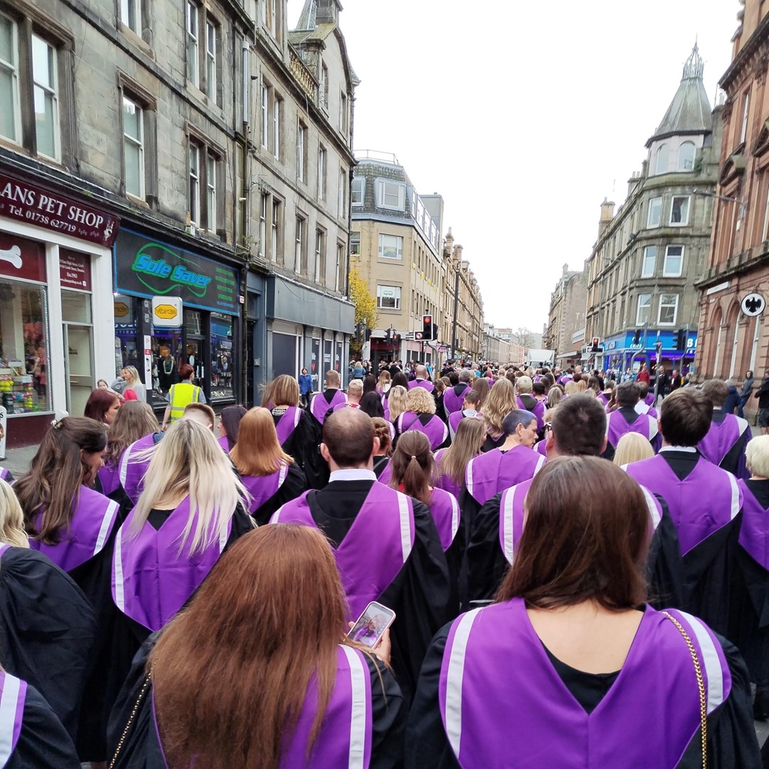 Group of people wearing graduation gowns and hoods, walking down a street with their backs to the camera