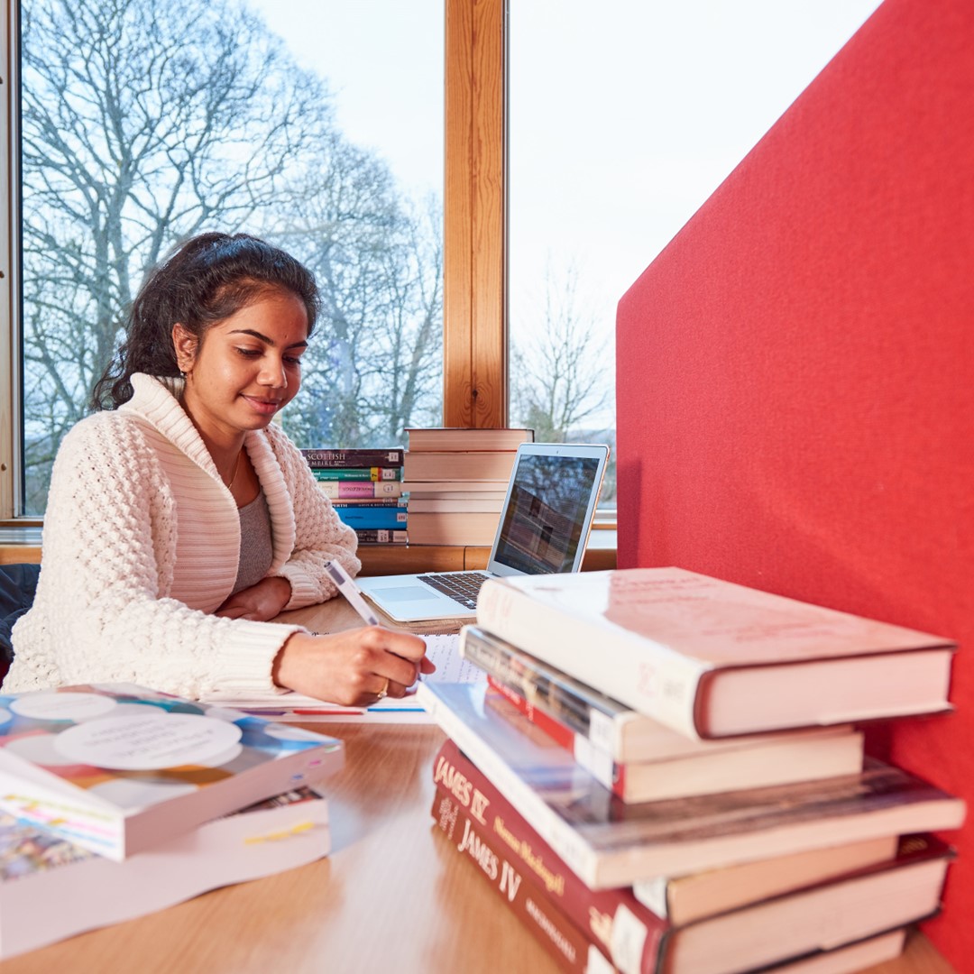 Woman taking notes at a desk, with a pile of books in front of her