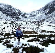 Woman wearing hiking gear, standing in a snowy landscape of hills and grassland. 