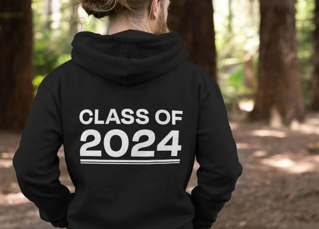 A man standing in the woods, with his back to the camera and his hoodie says 'Class of 2024